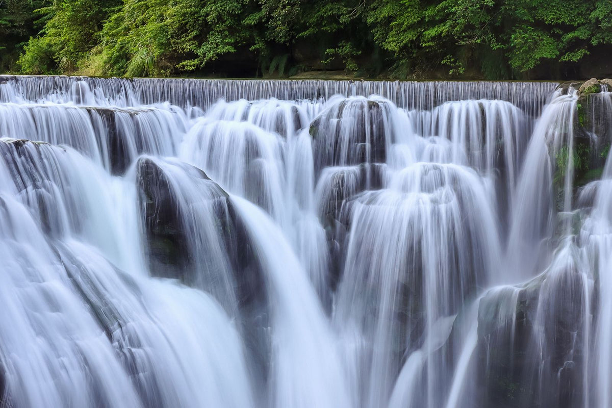 Brede waterval die in zachte stroken over rotsen valt, met groen bos erboven